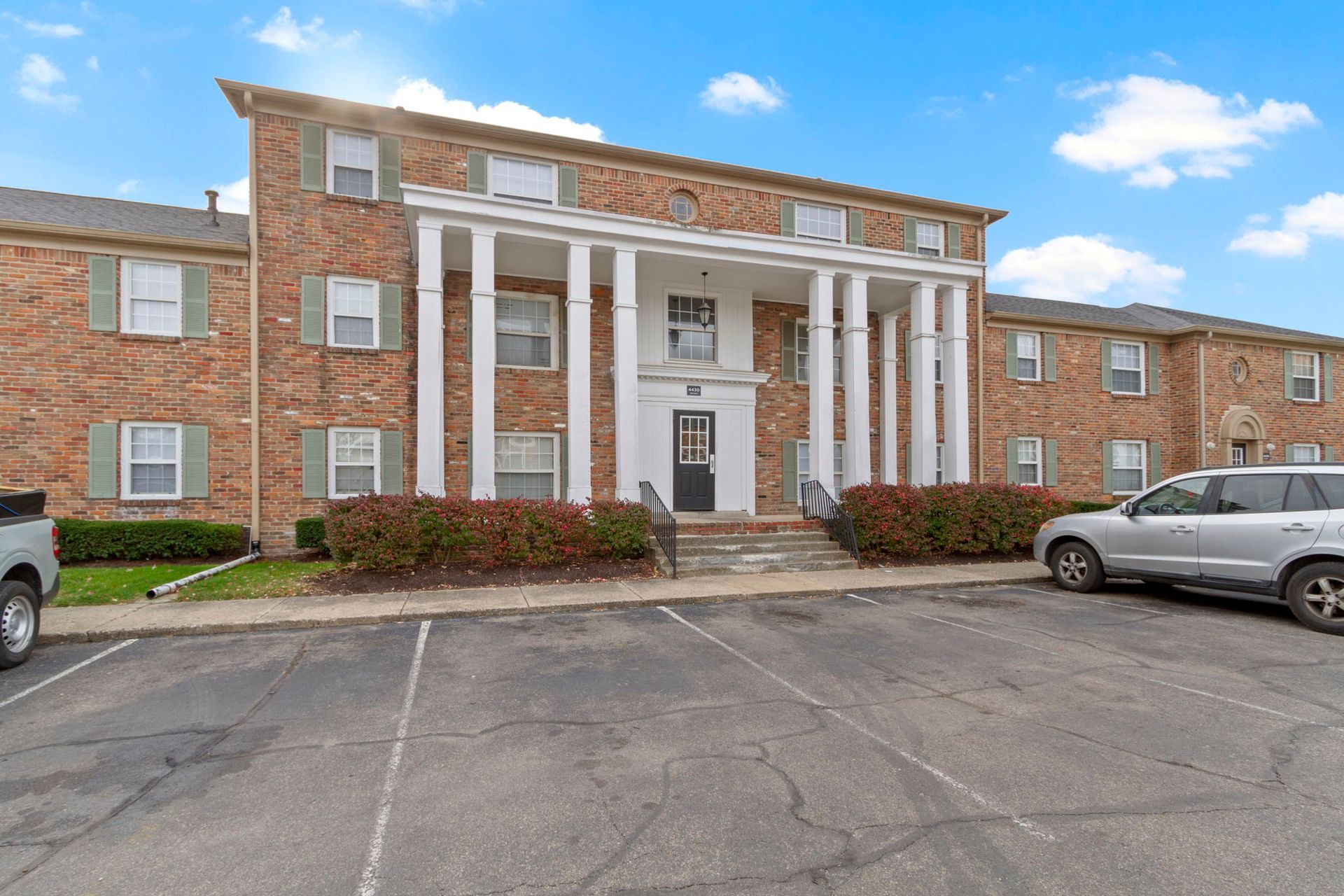 Apartment building with brick facade, white columns, and parking lot in front.