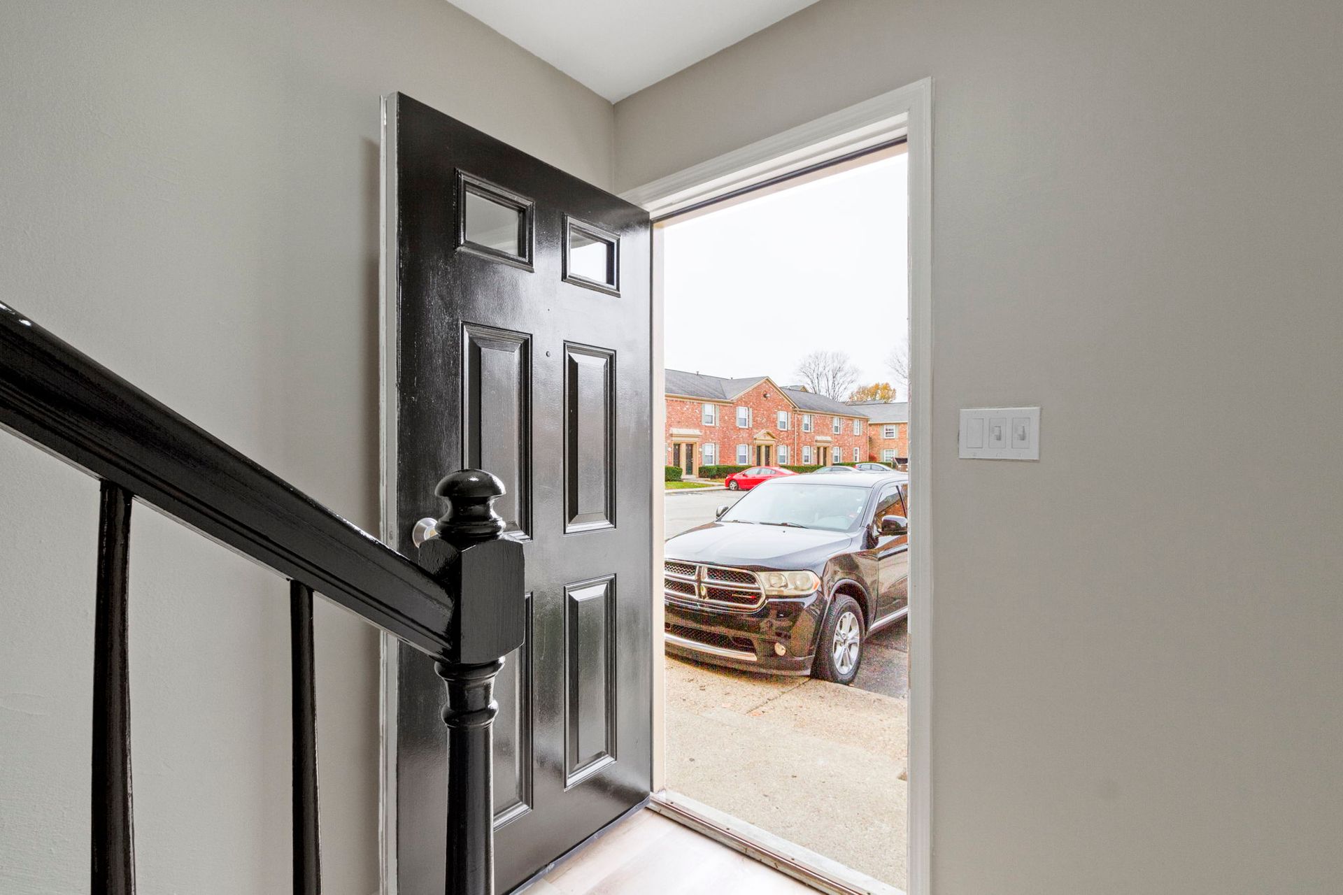 Black door open, revealing a car parked outside a brick building. Black stair railing on the left.