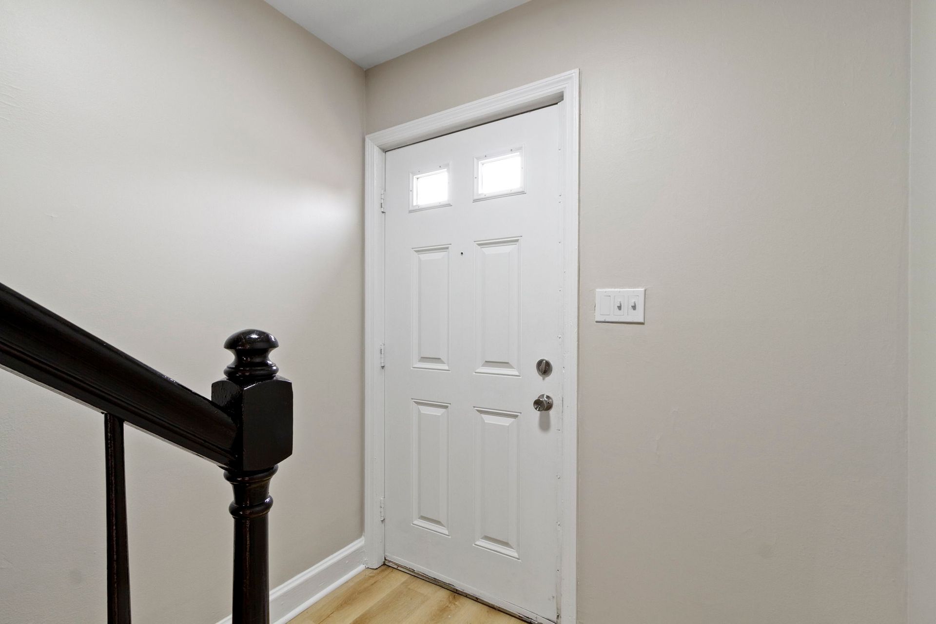 White door with small upper windows, beige walls, and a dark staircase.