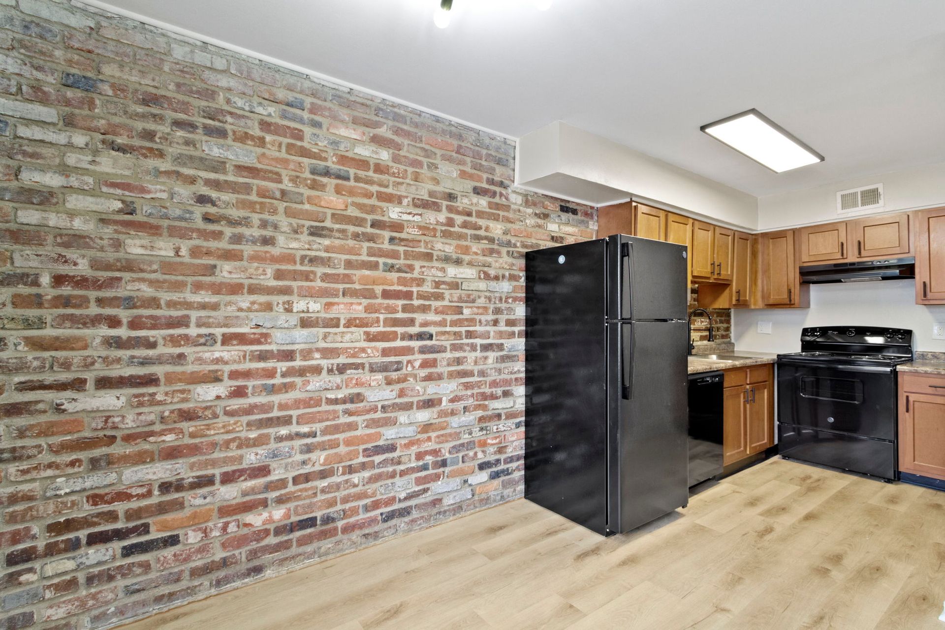 Kitchen with brick wall, black fridge, and wooden cabinets; light flooring.