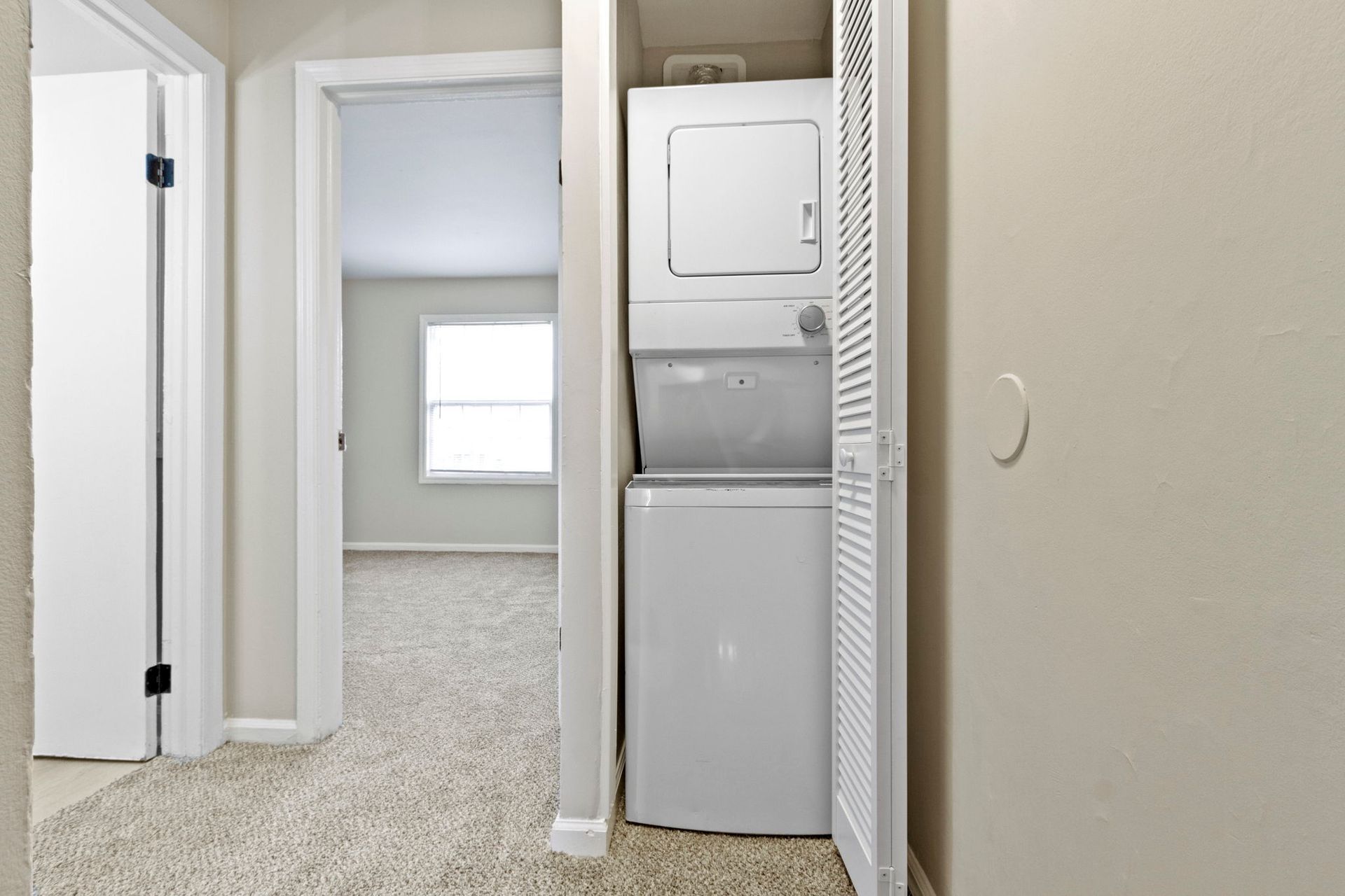 Hallway with stacked washer/dryer in closet, door on left, view of room with window.