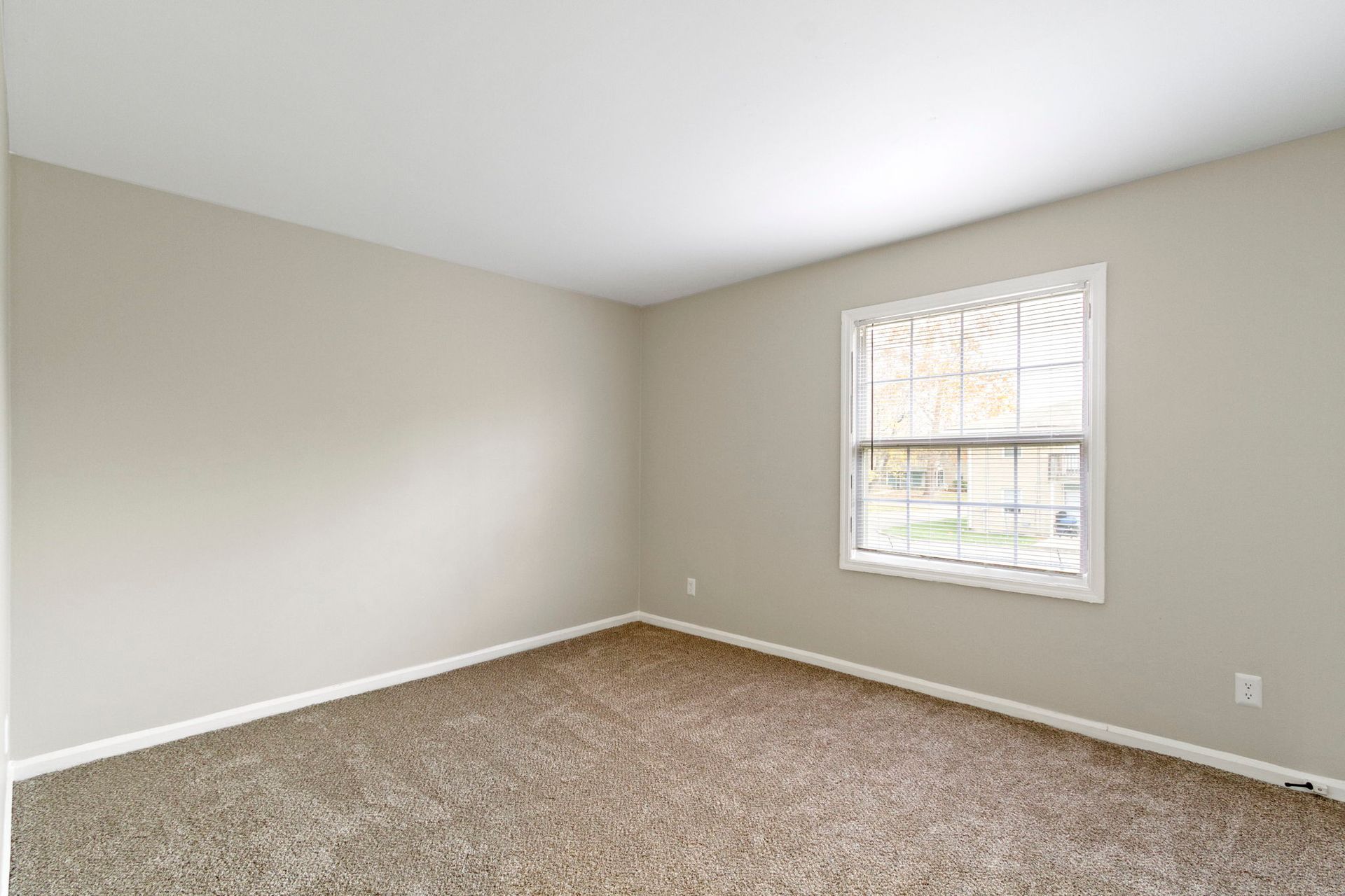 Empty bedroom with beige walls, carpet, and window with blinds.