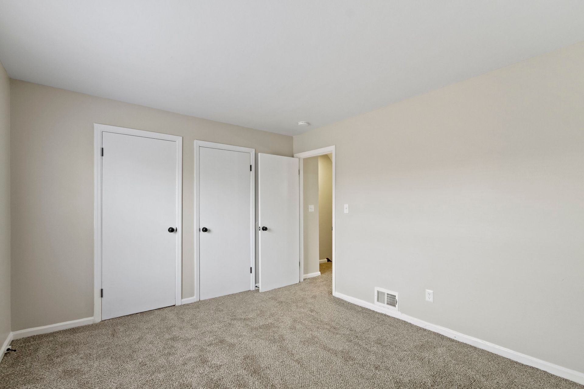 Empty bedroom with three white closet doors, carpet, and beige walls.