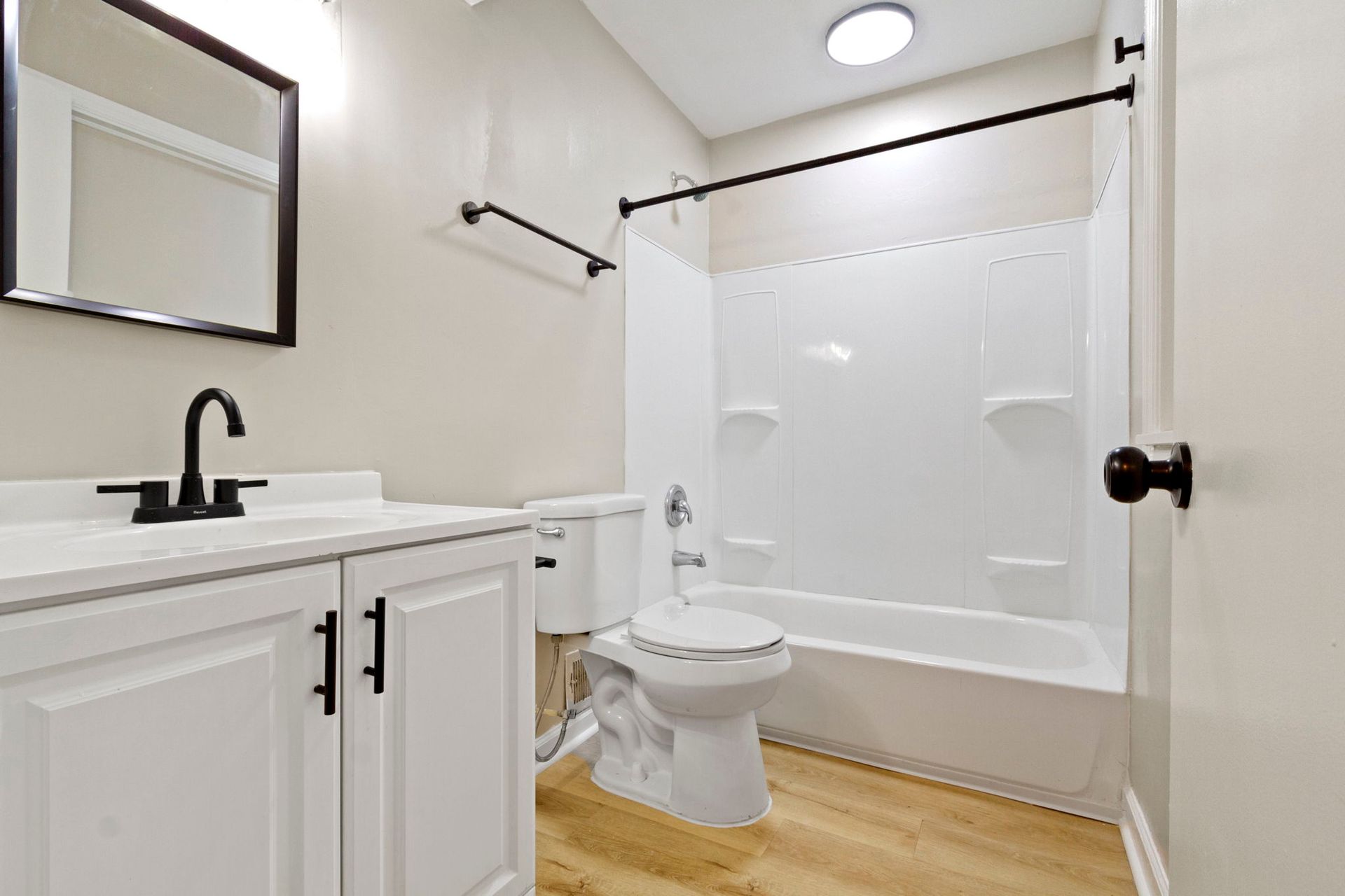 White bathroom with white vanity, toilet, and tub, black accents, and light wood-look floor.