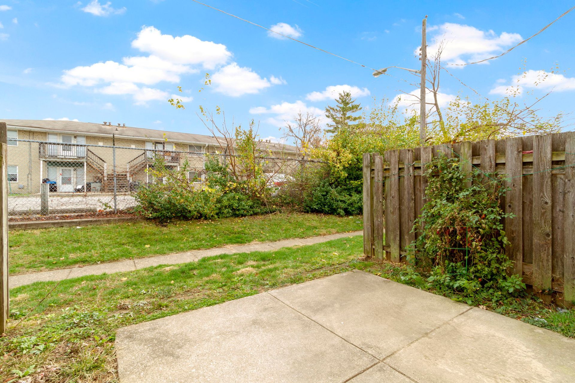 Patio and backyard with green grass, a wooden fence, and buildings in the background under a blue sky.