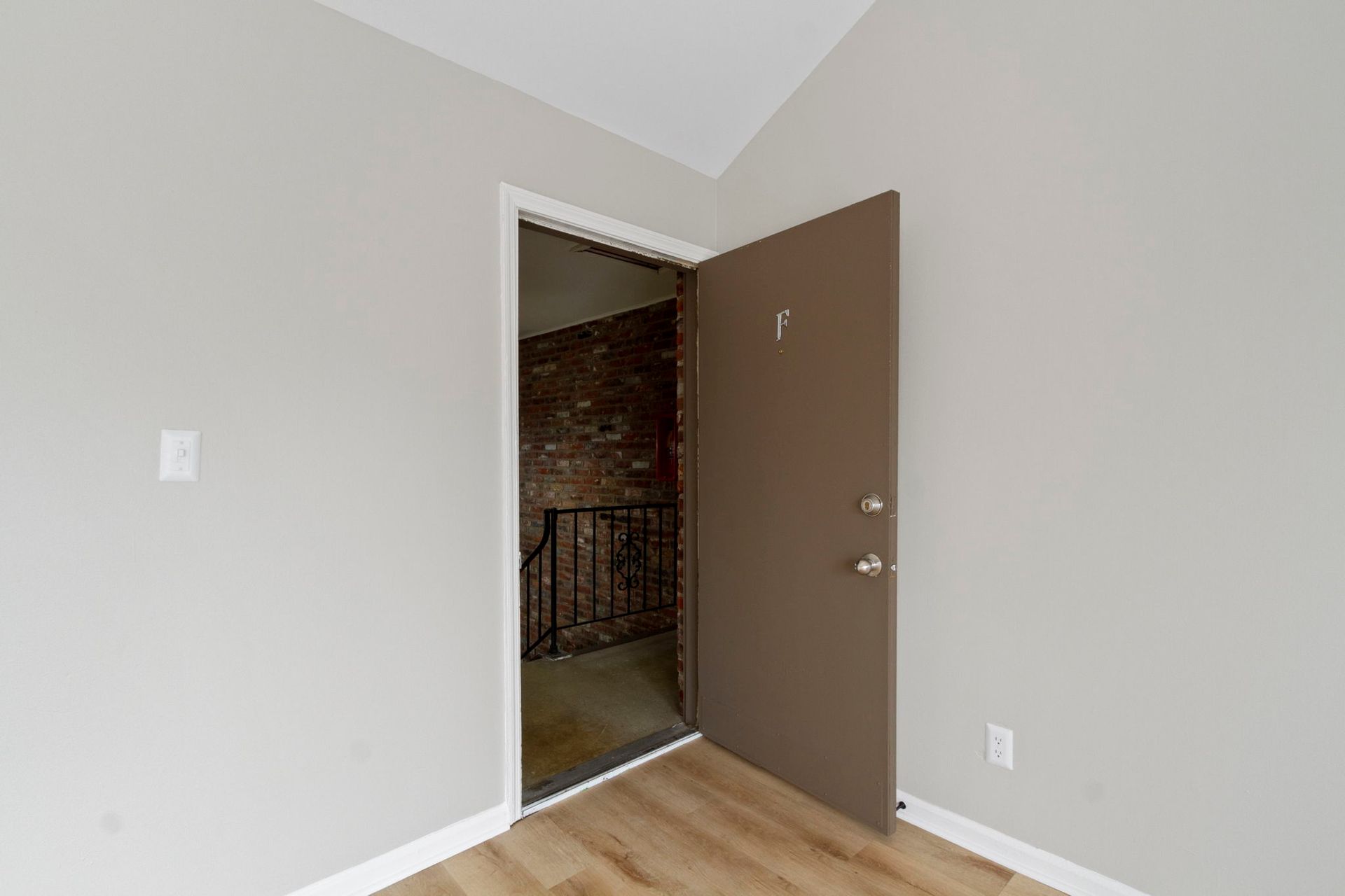 Brown door open to a stairwell with brick wall and black railing. Light gray walls and wood-look floor.