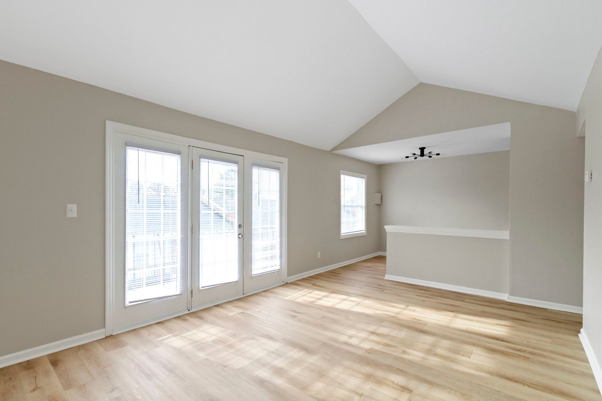 Empty living room with light wood floors, tan walls, and a vaulted ceiling. French doors and windows provide natural light.