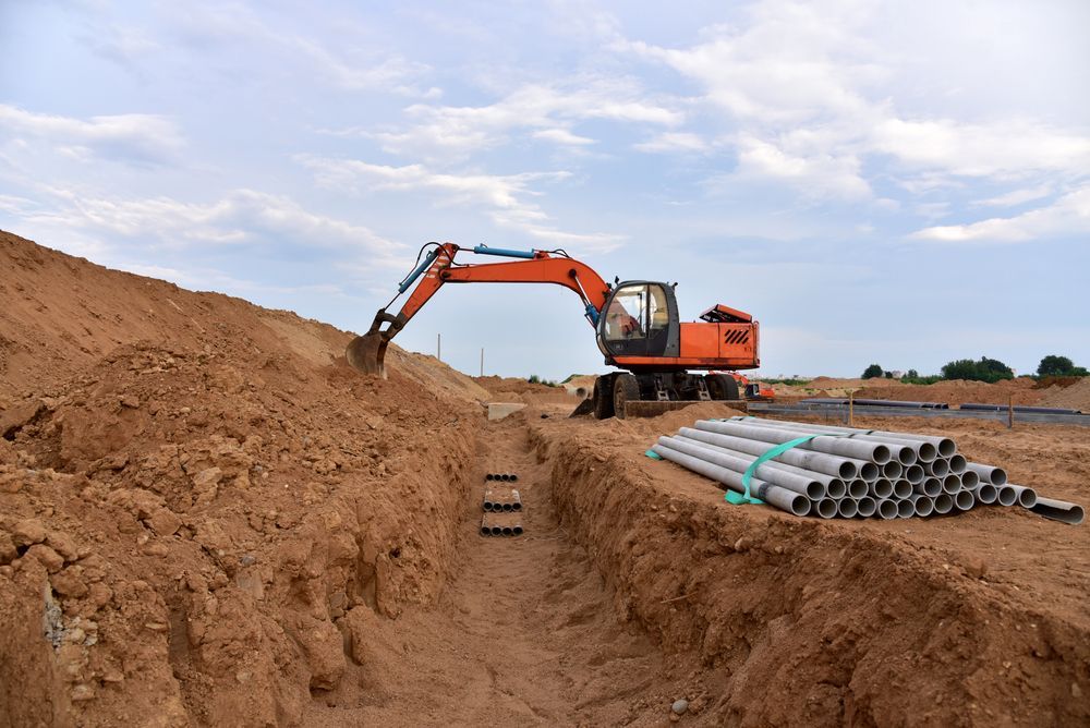 An orange excavator digs a trench on a construction site, with a pile of concrete pipes nearby. Cloudy sky.
