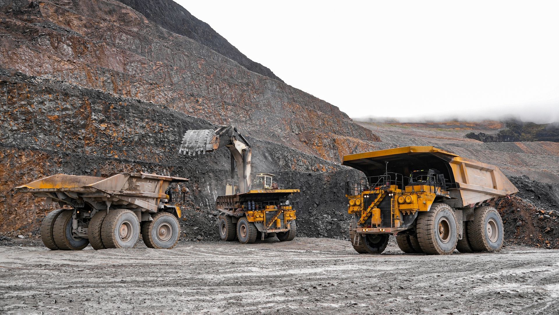 Yellow mining trucks and an excavator at a mine site, moving ore against a gray sky.