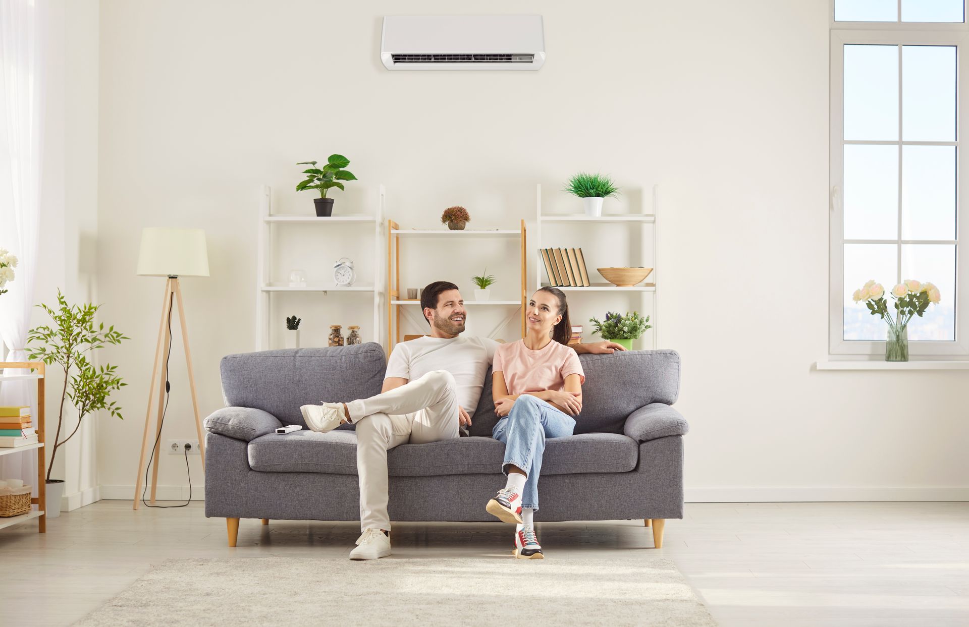 Couple relaxing on a gray sofa in a bright living room with white shelves and plants. Couple relaxing on a gray sofa in a bright living room with white shelves and plants.