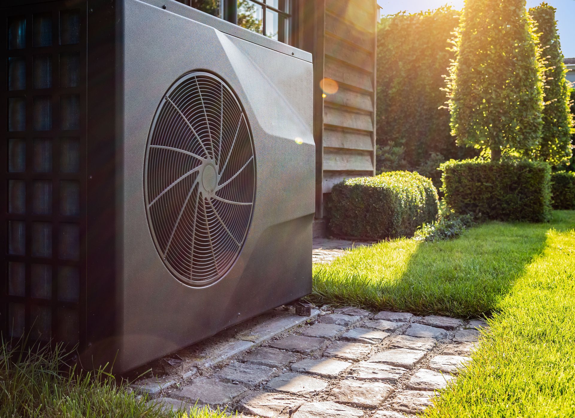 Outdoor air conditioner unit beside a stone path, with sunlight and green grass in the yard