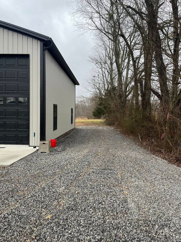 Gravel driveway beside a gray metal building, with trees and shrubs along the right side.