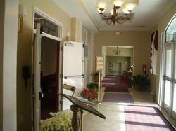 Hallway with open doors leading to a chapel and a further room, flowers, burgundy carpet, beige walls, and a chandelier.
