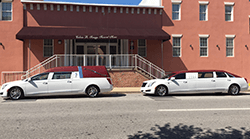 White hearse and limousine parked in front of a brick building with a funeral home sign.