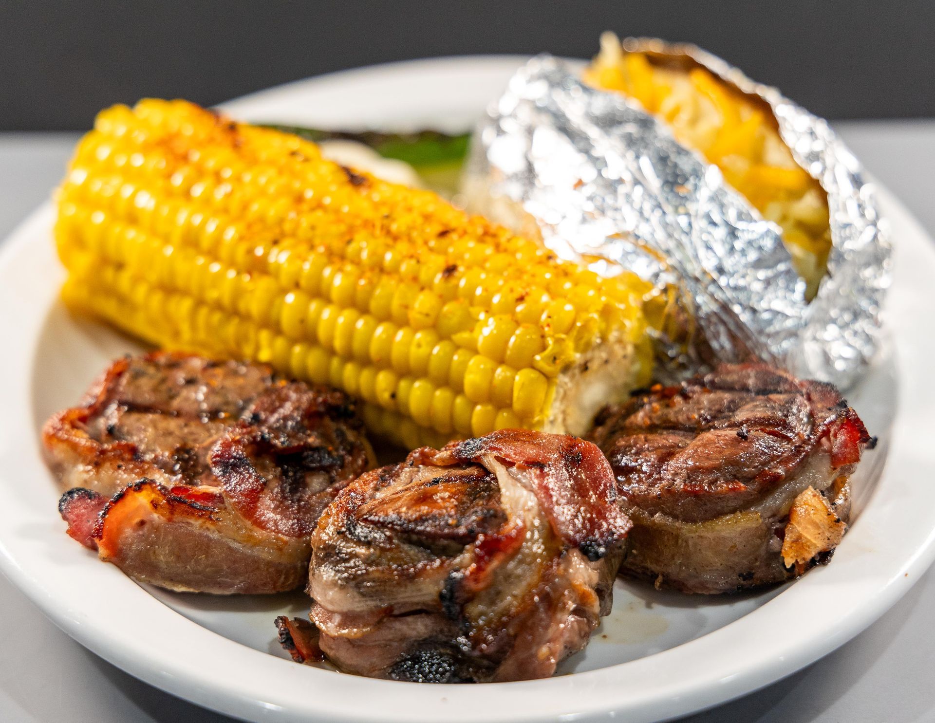 A white plate topped with meat , corn on the cob and baked potatoes.