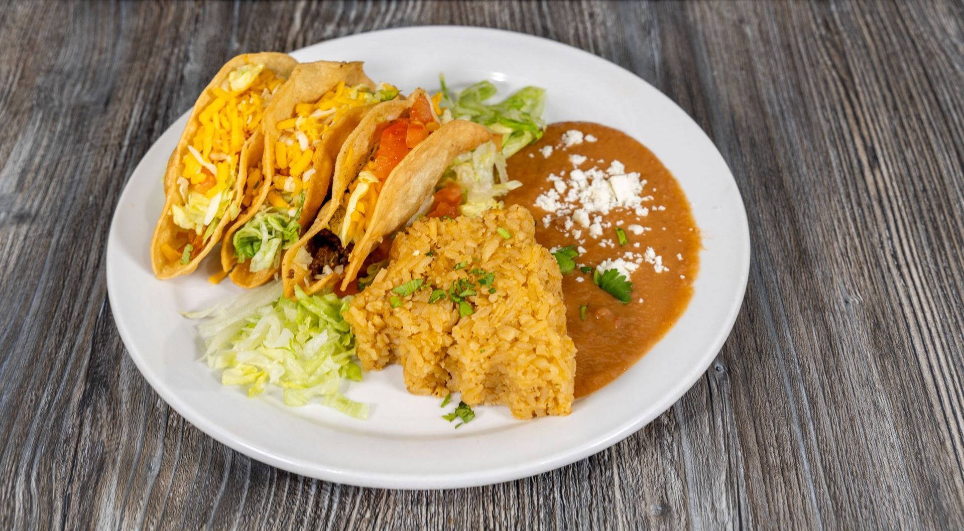 A white plate topped with tacos , rice and beans on a wooden table.