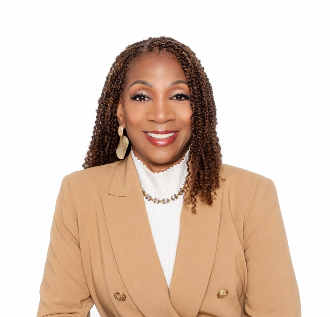 Woman with curly brown hair, smiling, wearing a pink top, black jacket, and gold necklace.