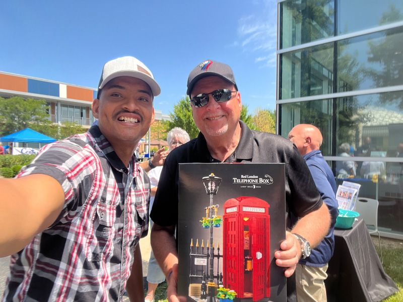 Two men smiling for a selfie; one holds a framed London phone booth art print; outdoor setting.