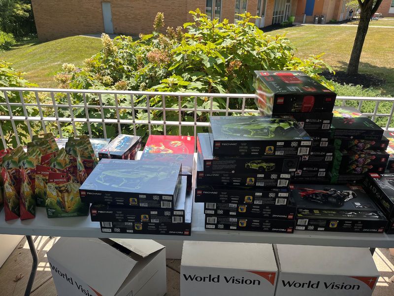 Table with stacks of Lego sets and gift bags, outdoors. Boxes are black, red, and blue. World Vision boxes are in the front.