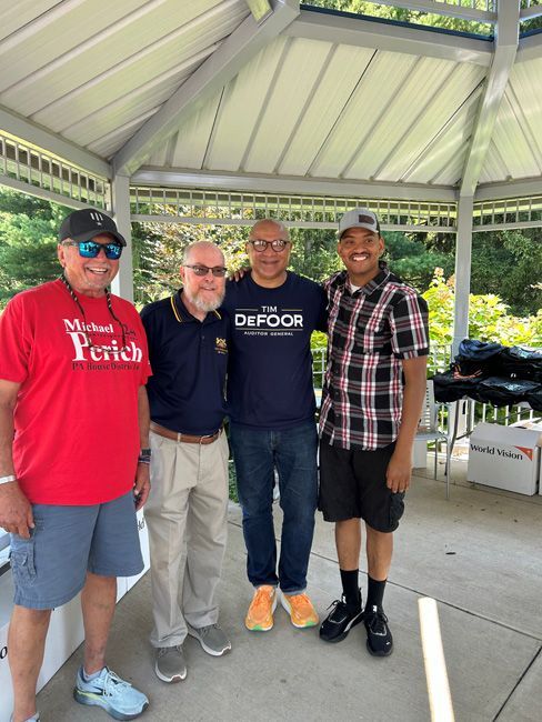 Four men standing together under a gazebo; two are smiling. One wears a red shirt that says 