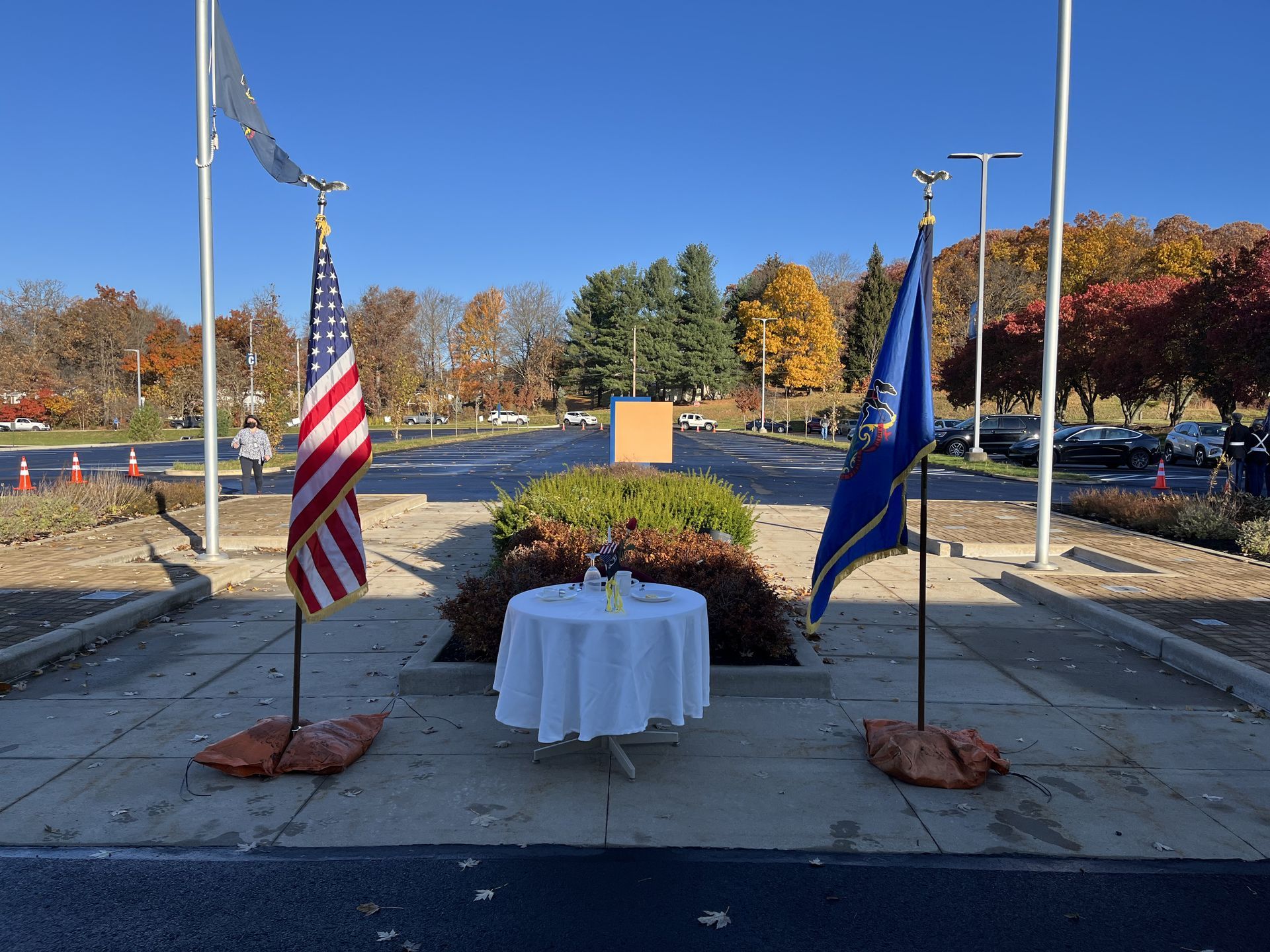 American and state flags flank a table with a white cloth, set in front of a monument. Autumn trees surround.
