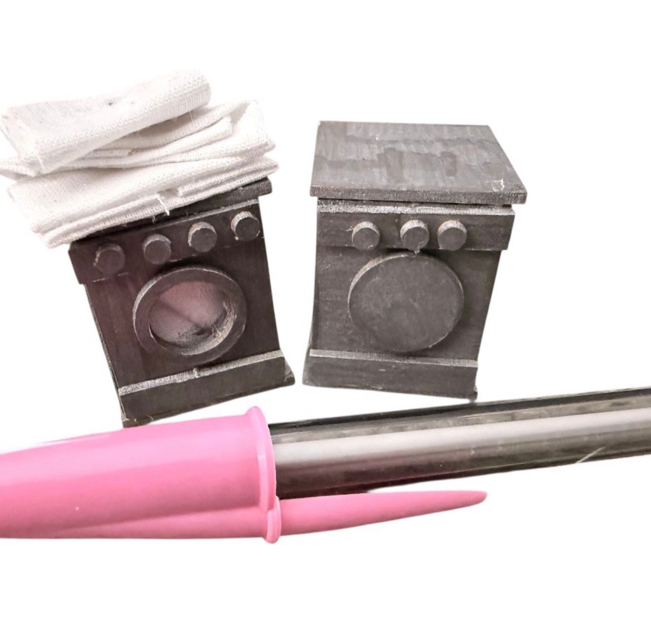 Two gray toy washing machines with a pile of white fabric and a pink tool.