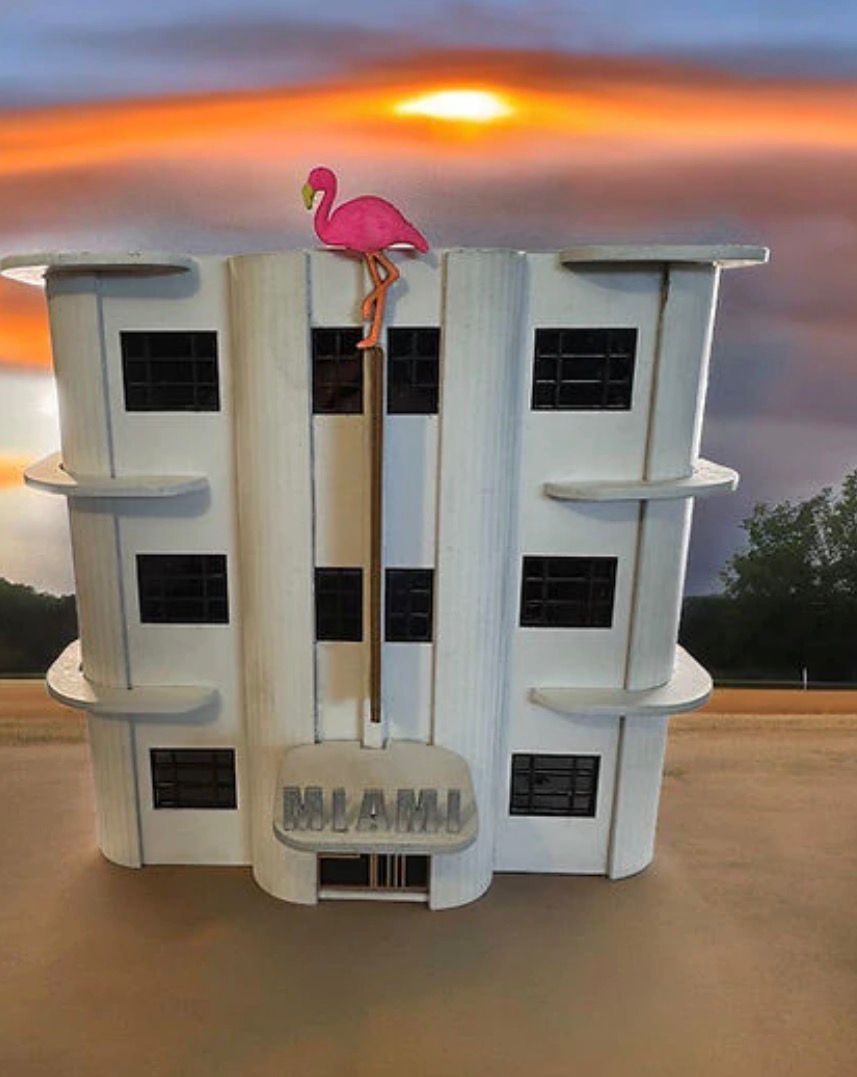 Model of a white Art Deco building with a pink flamingo on top, against an orange sunset.