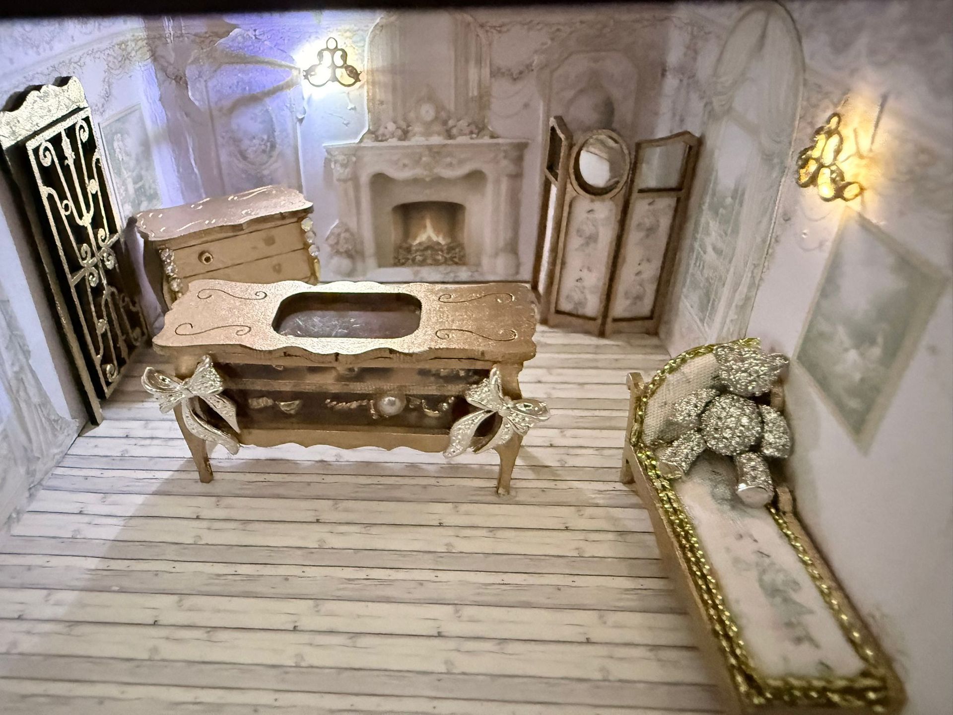 Miniature gold and white room: dresser, fireplace, screen, bed with bejeweled teddy bear, and sconces.