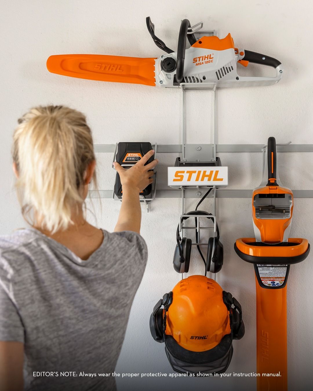 Woman hanging battery on a STIHL wall storage system. Chainsaw, accessories, and gear are mounted on a wall.