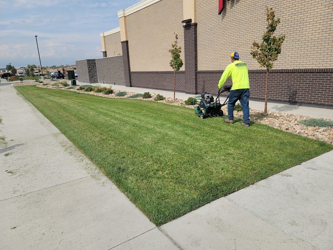 A worker in a neon yellow shirt uses a walk-behind mower to trim a grassy strip alongside a building and sidewalk.