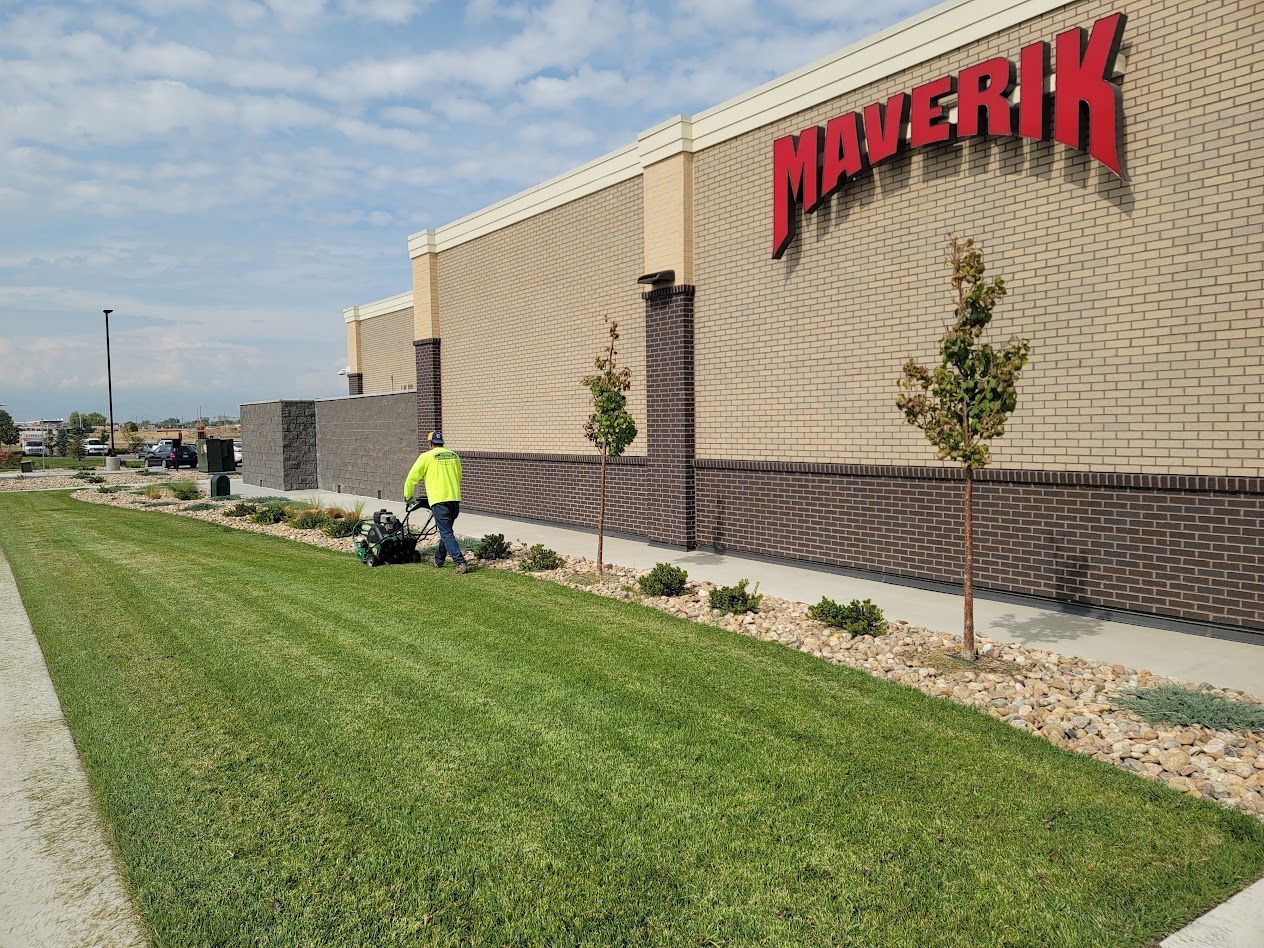 A person in a bright yellow shirt mows the grass in front of a tan brick Maverik building under a blue, cloudy sky.