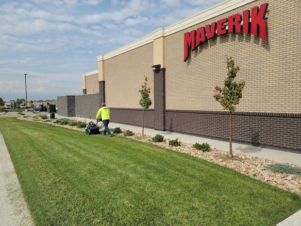 A person in a bright yellow shirt mows the grass in front of a tan brick Maverik building under a blue, cloudy sky.