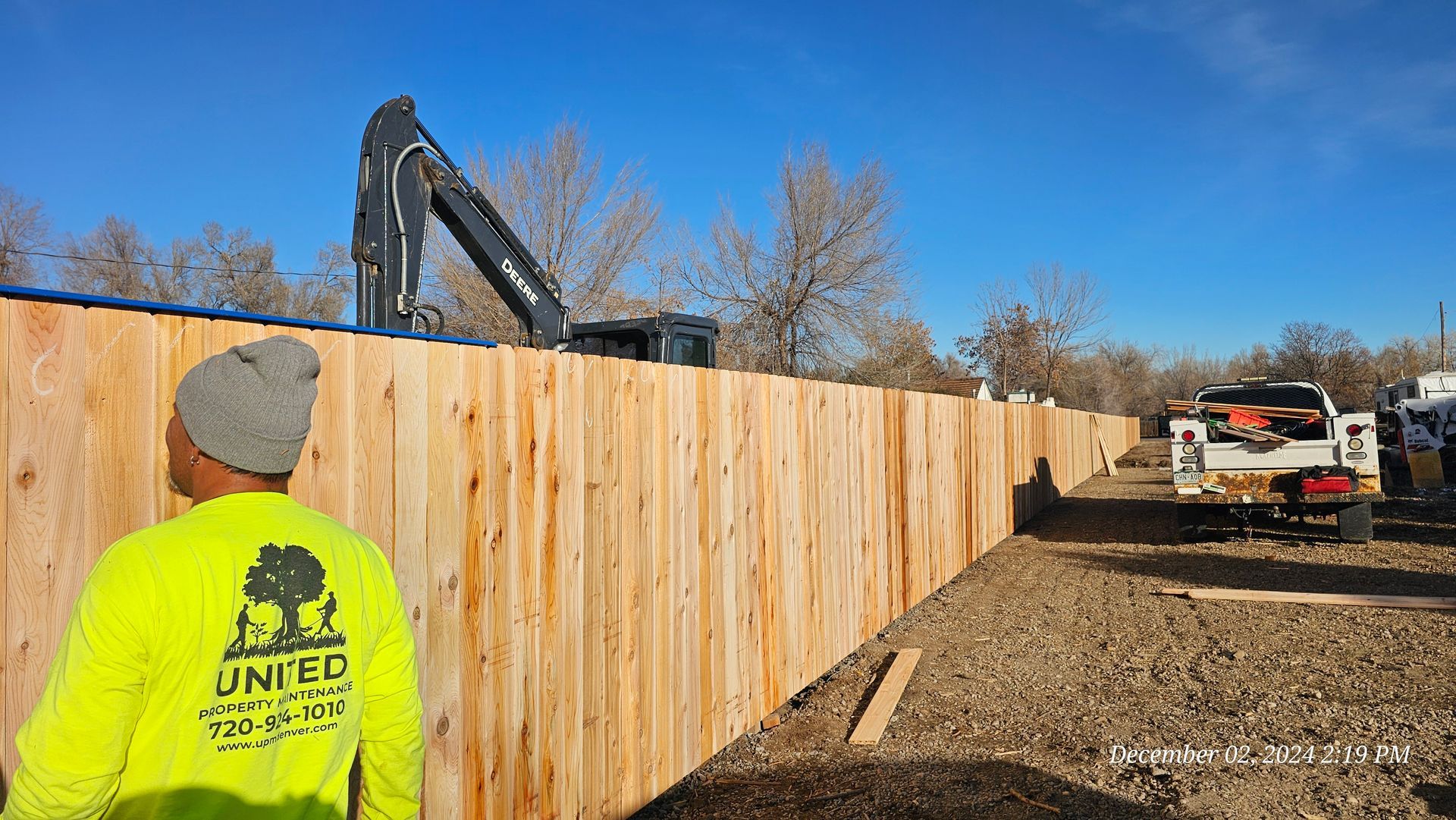 A worker in a bright yellow long-sleeved shirt stands before a long wooden fence at an outdoor construction site.