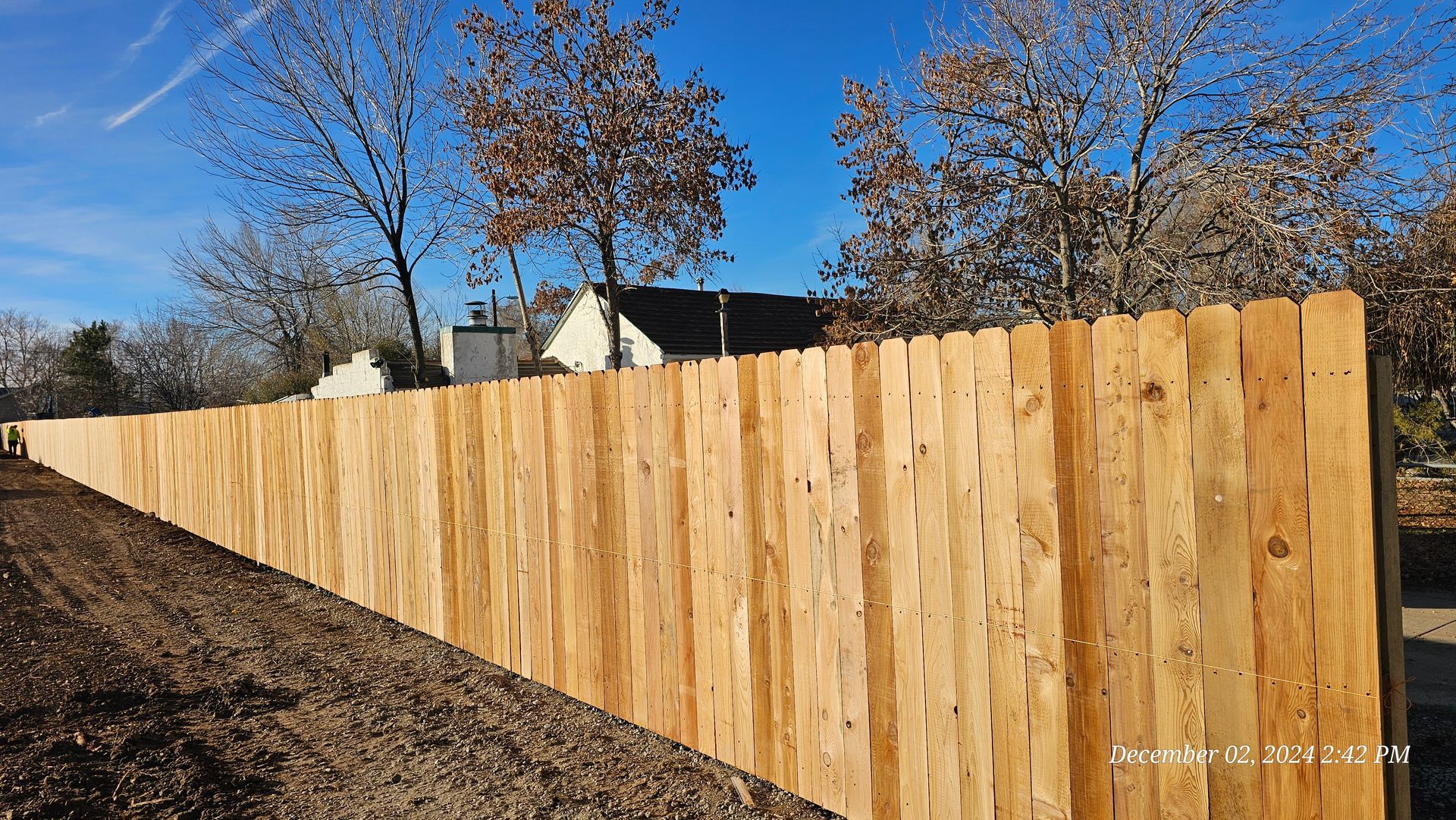 A newly installed wooden picket fence stands alongside a dirt path under a clear blue sky with trees in the background.