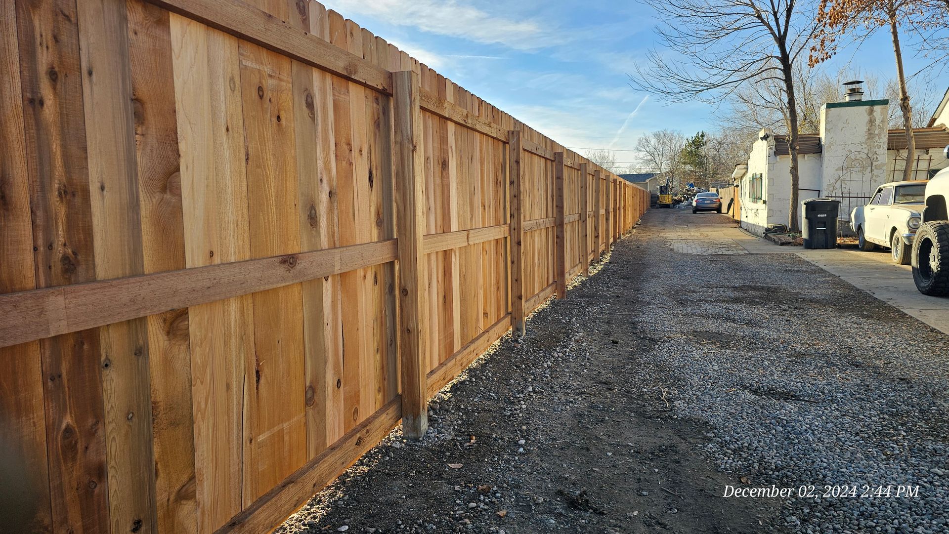 A long, newly installed wooden privacy fence runs alongside a gravel path leading toward buildings in the distance.
