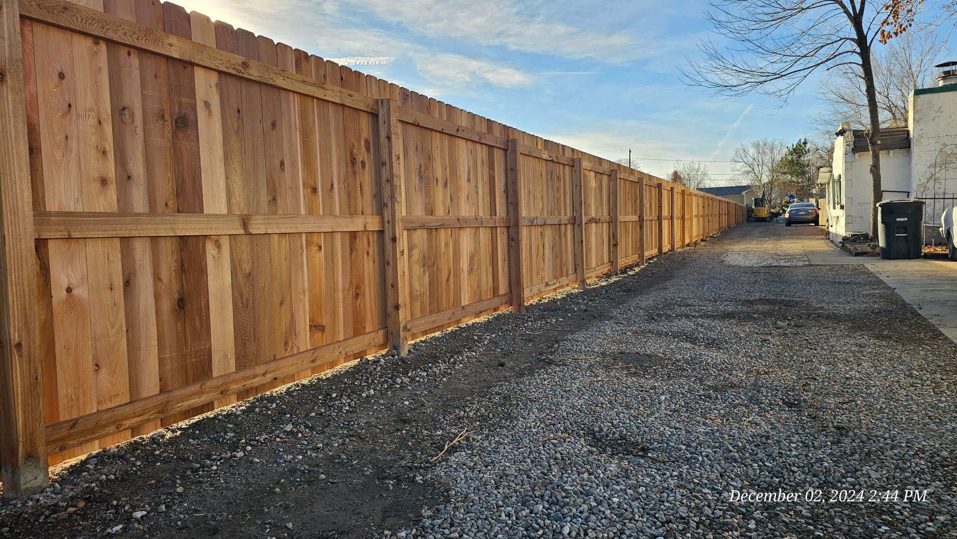 A long, new wooden privacy fence stretches along a gravel-covered pathway outdoors under a clear blue sky.