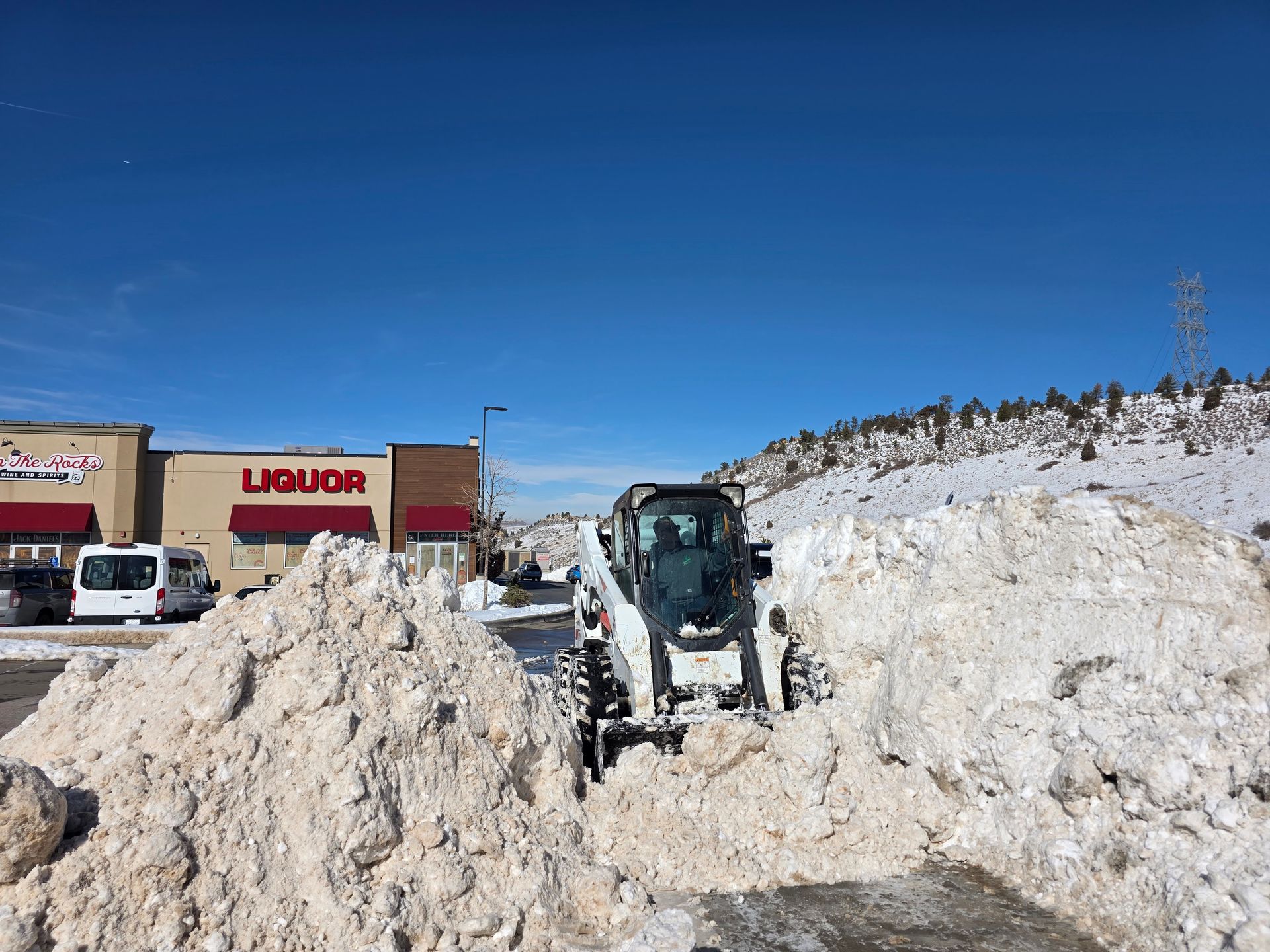 A white skid steer loader clears deep snow in a commercial parking lot near a liquor store under a clear blue sky.