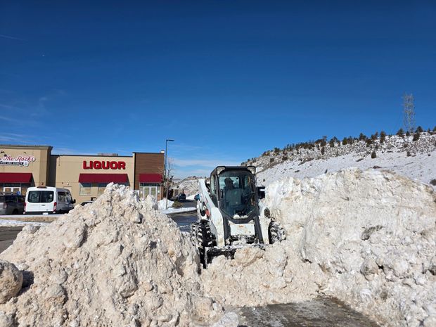 A white skid steer loader clears deep snow in a commercial parking lot near a liquor store under a clear blue sky.