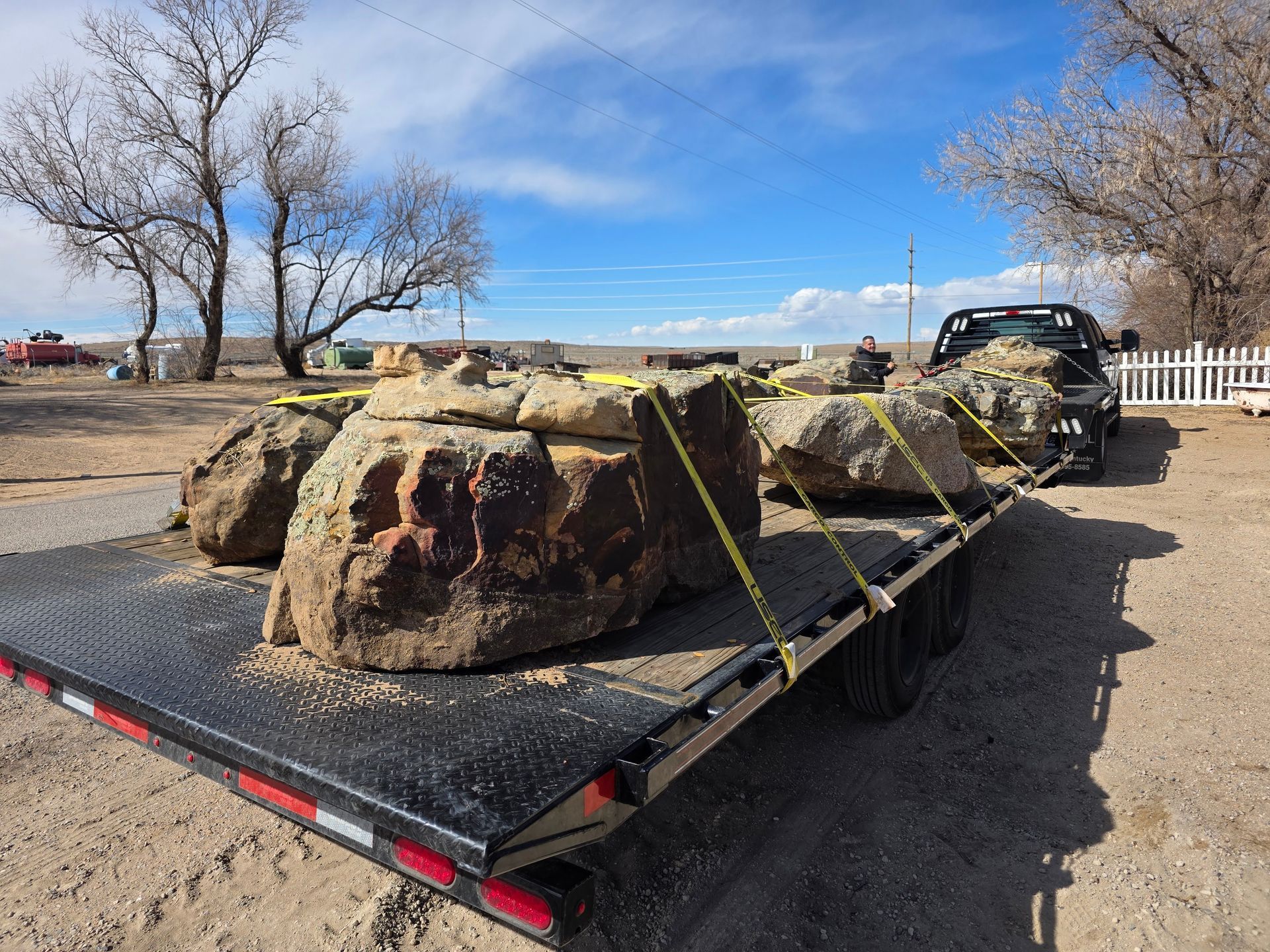 A flatbed trailer loaded with large, irregular brown boulders secured by yellow straps, parked on a dirt lot.