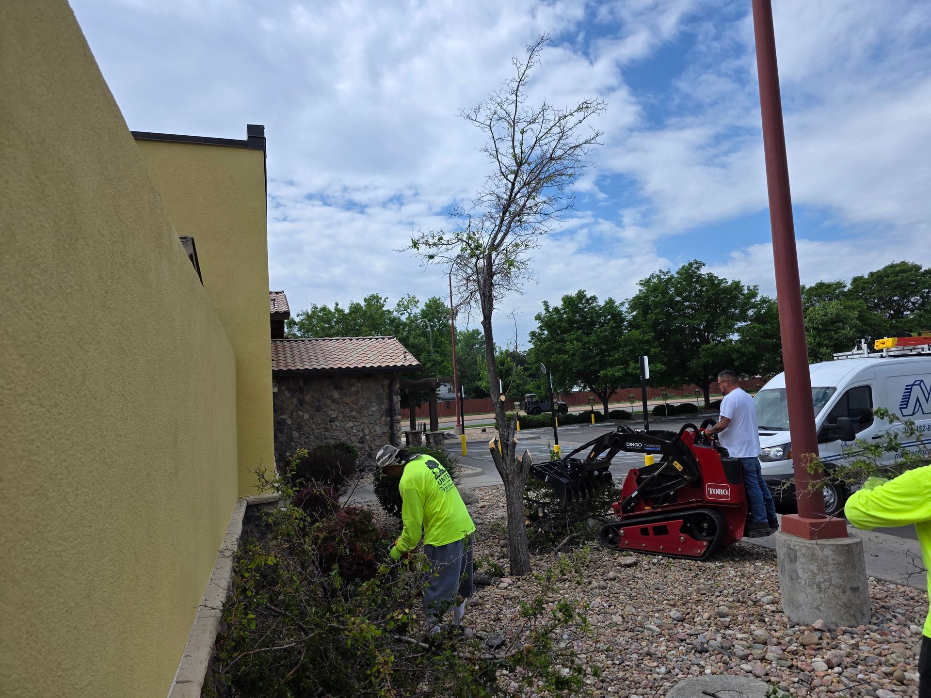 Two workers in neon-yellow shirts landscape a rocky garden bed next to a yellow building with a small tractor.
