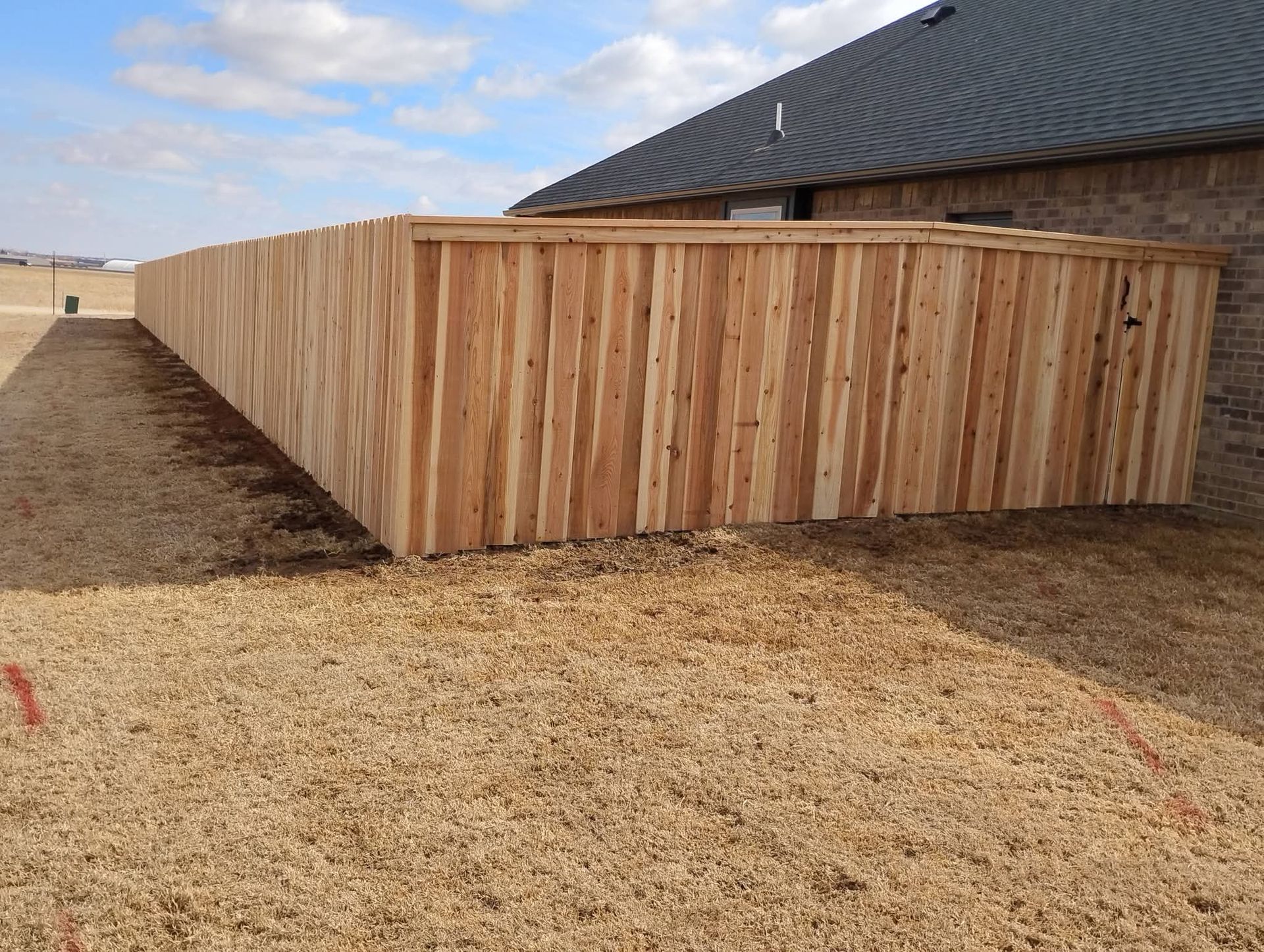 A side view of a tall, newly installed wooden privacy fence running alongside a brick house on a dirt lot.