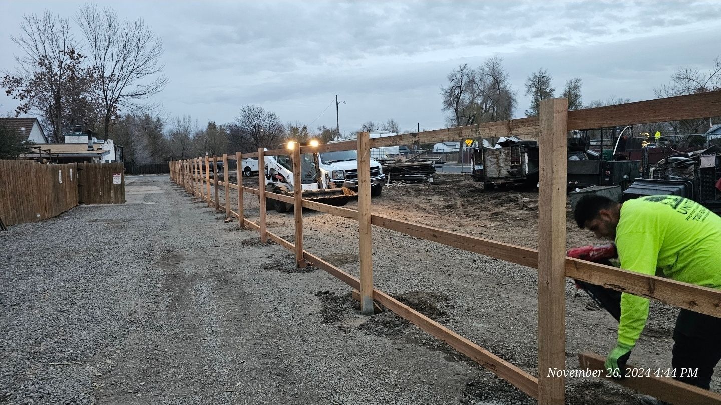 A worker in a neon green shirt builds a wooden post-and-rail fence at a gravel construction site.
