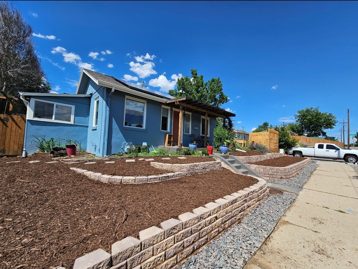 A blue house with a terraced front yard featuring stone retaining walls, fresh mulch, and a sidewalk on a sunny day.