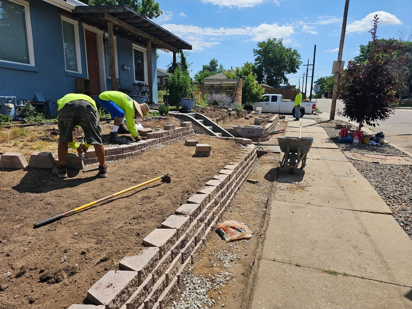 Two people in high-visibility yellow shirts perform landscaping work in the front yard of a blue house beside a sidewalk.