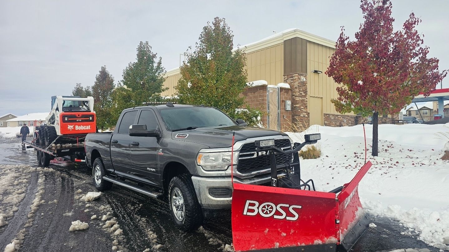 A gray pickup truck with a red BOSS snowplow attached to the front, towing a Bobcat on a trailer in a snowy parking lot.