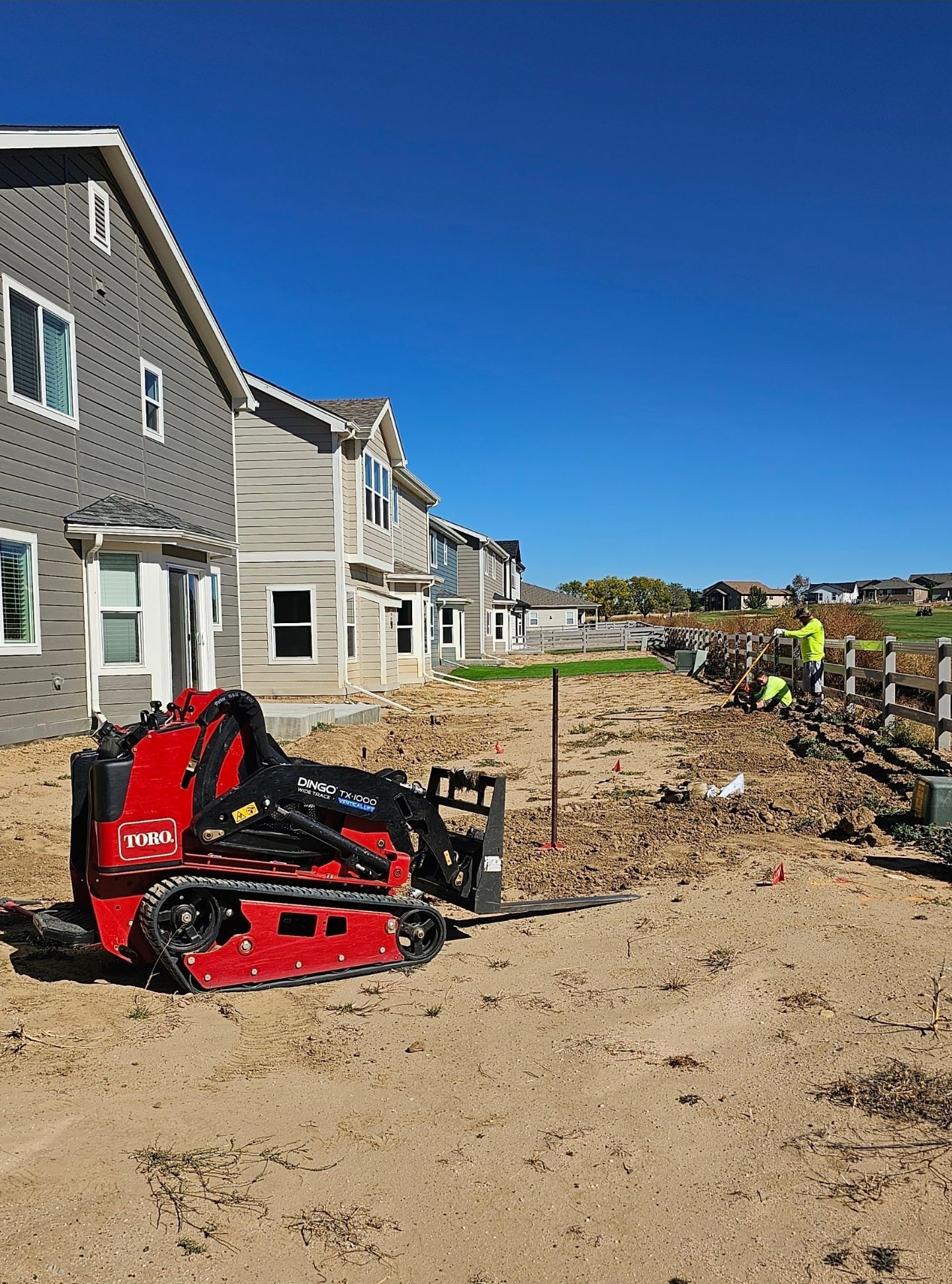 A red Toro tracked skid steer parked on a dirt lot next to a row of houses under construction under a clear blue sky.