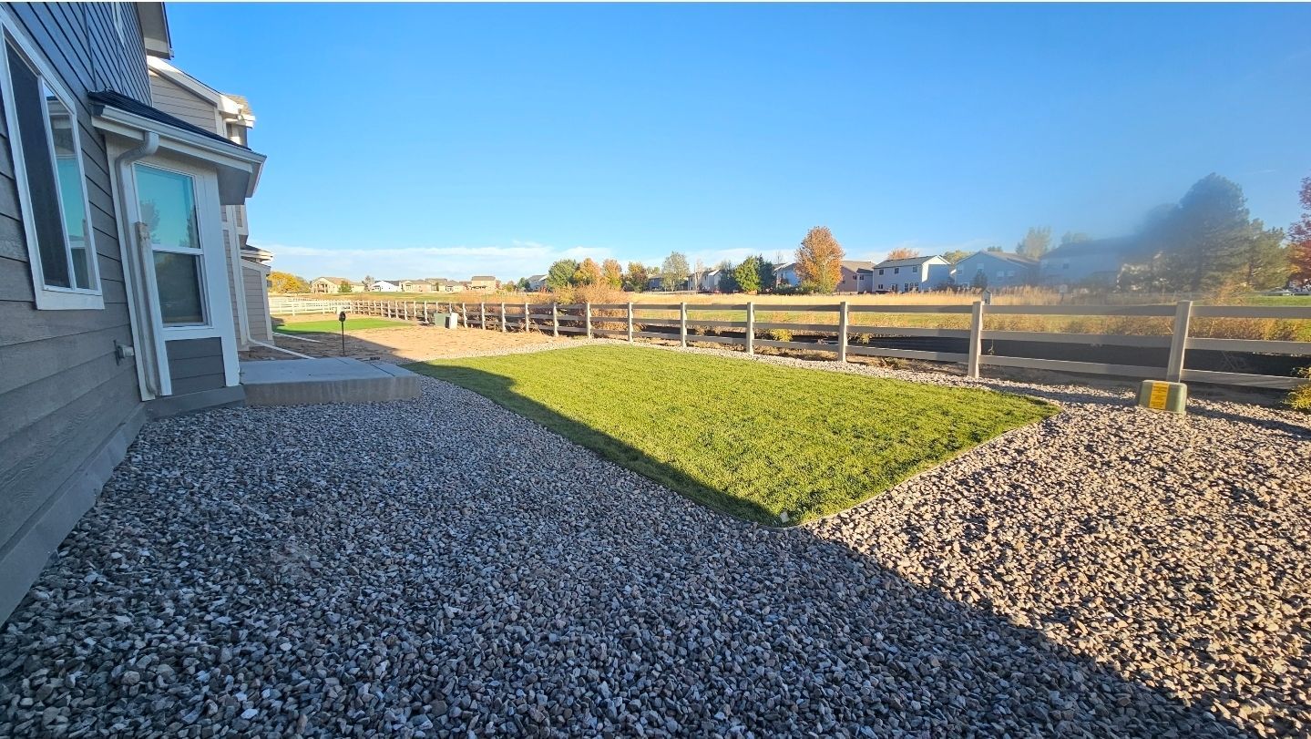 A side yard with a rectangular patch of green grass surrounded by light-colored gravel, next to a grey home on a sunny day.