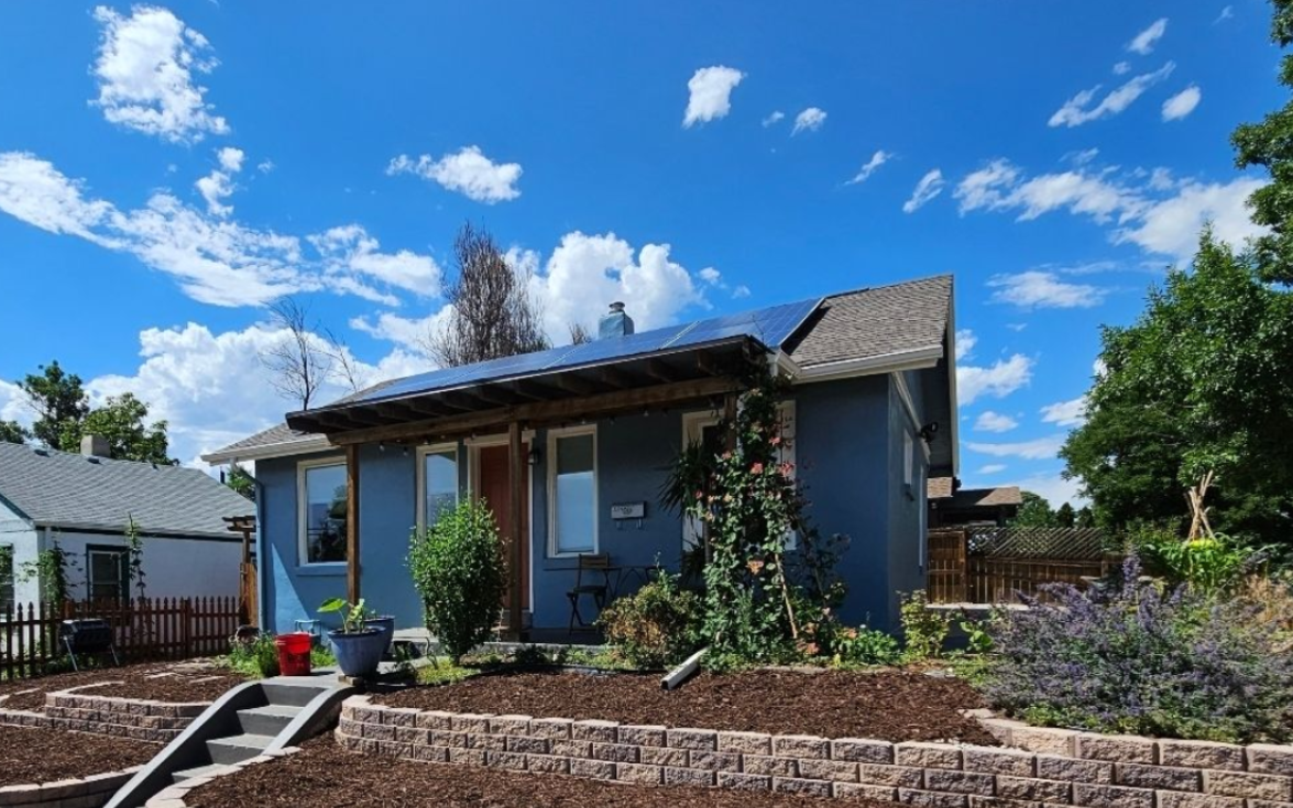 A blue house with solar panels on the roof, surrounded by landscaping, terraced stone retaining walls, and a blue sky.