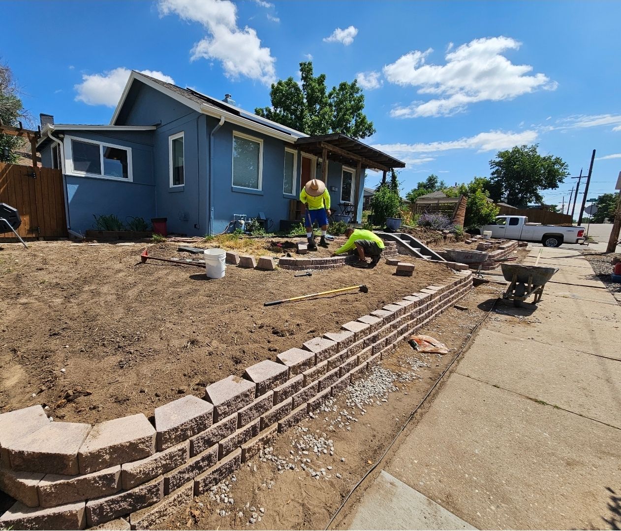 Two workers in safety vests install a decorative brick retaining wall in the front yard of a blue house under a sunny sky.