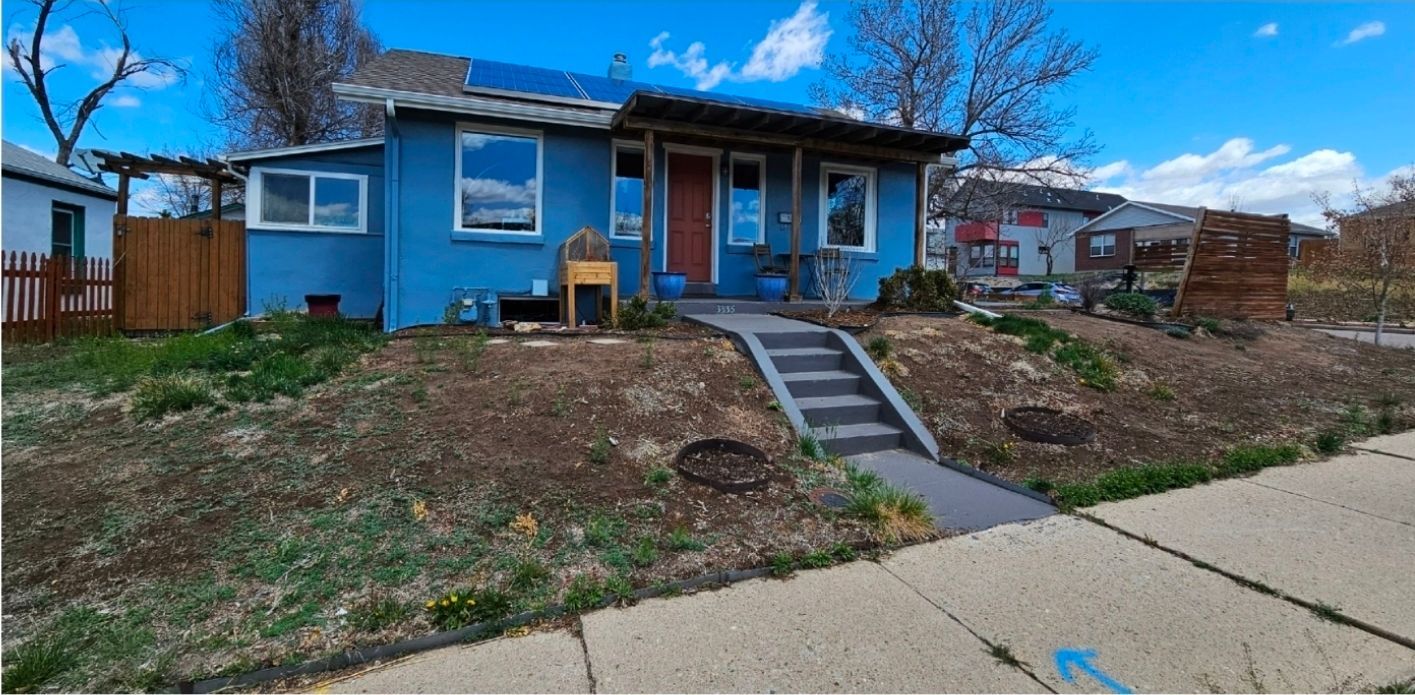 A bright blue, single-story house with a wooden porch, front steps, and solar panels sits on a sloped, grassy front yard.